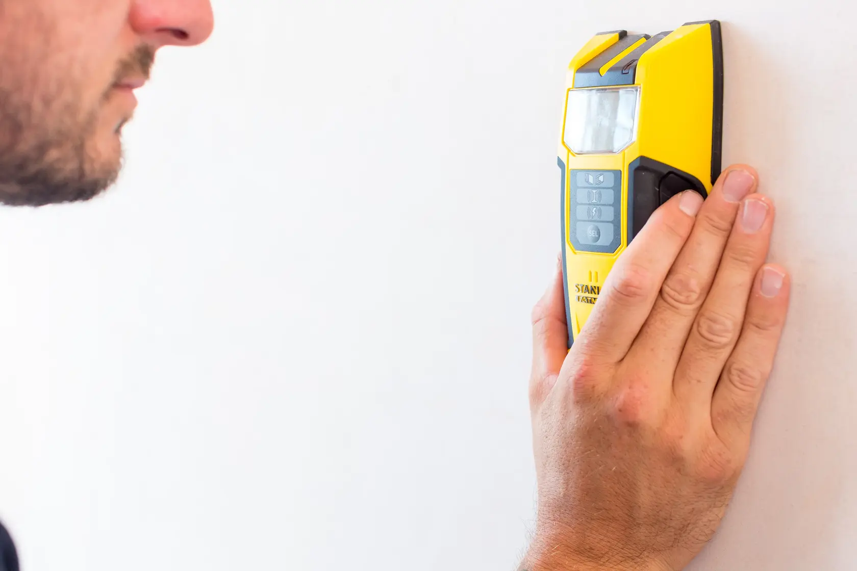 STANLEY FMHT0-77407 localizador electrónico amarillo sostenido en una pared blanca por una persona.