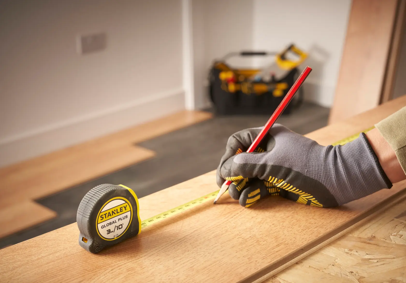 Fotografía de cinta métrica STANLEY 30-710 sobre tablón de madera, trabajador marcando medida, bolsa de herramientas al fondo.