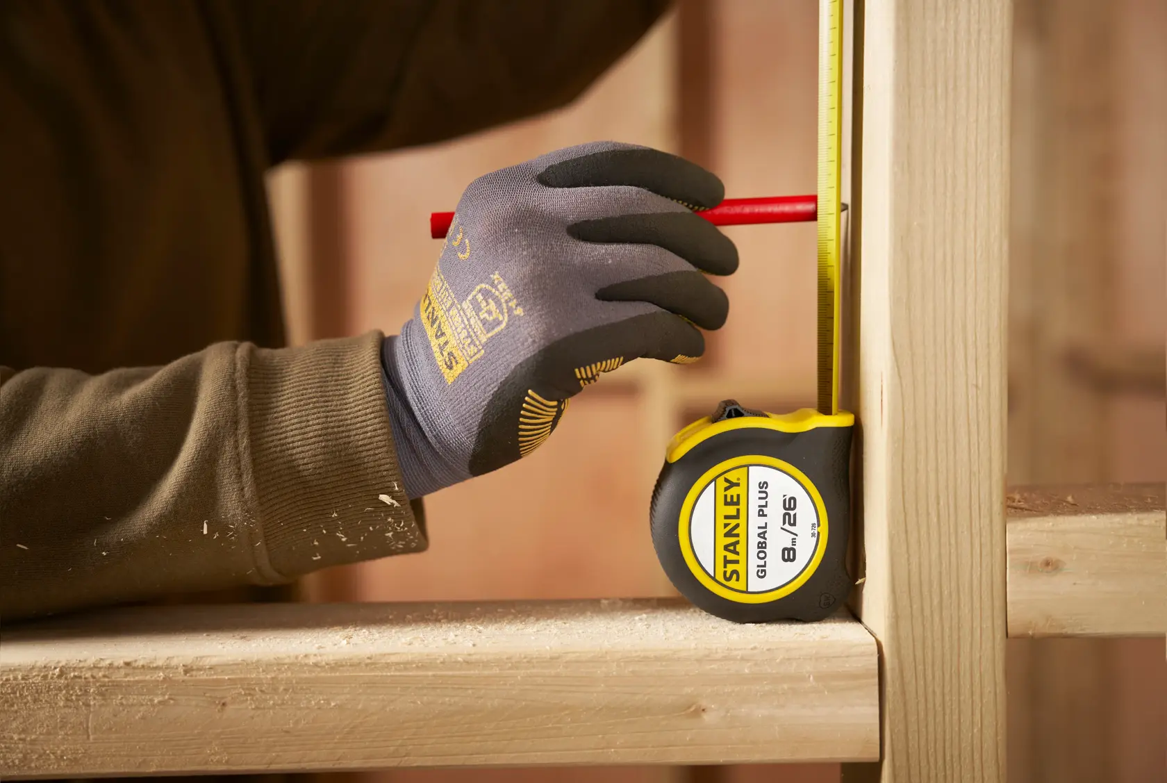 Cinta métrica STANLEY 30-726 utilizada para marcar madera, mano enguantada sujetando lápiz en tabla vertical en taller.