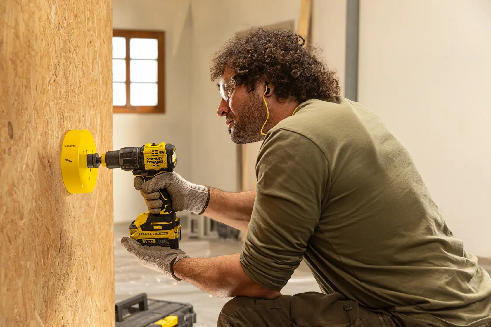 Persona utilizando un taladro inalámbrico STANLEY con una sierra de corona grande y amarilla para perforar un panel de madera. La persona lleva guantes y protección auditiva en un taller.