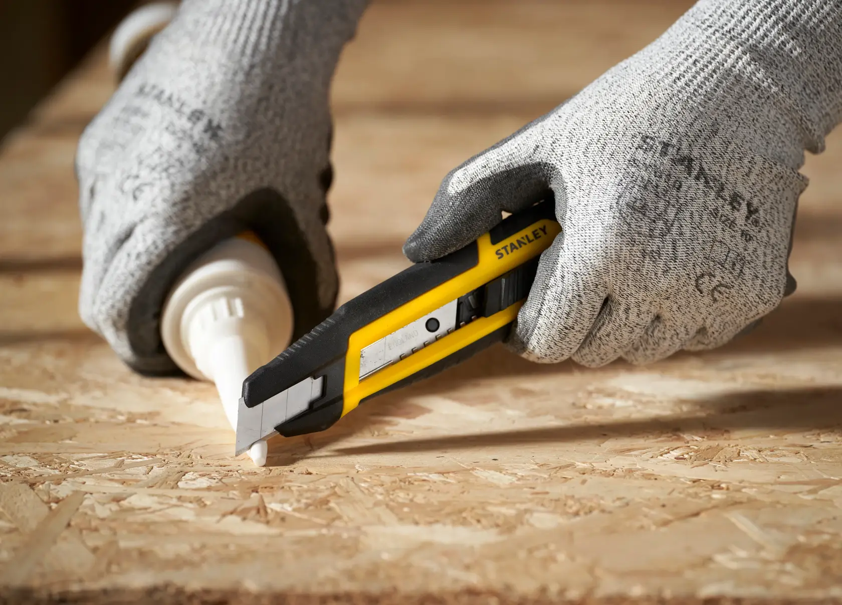 Fotografía de cuchillo multiusos STANLEY STHT10501 cortando madera, manos con guantes sostienen la herramienta amarilla y negra.