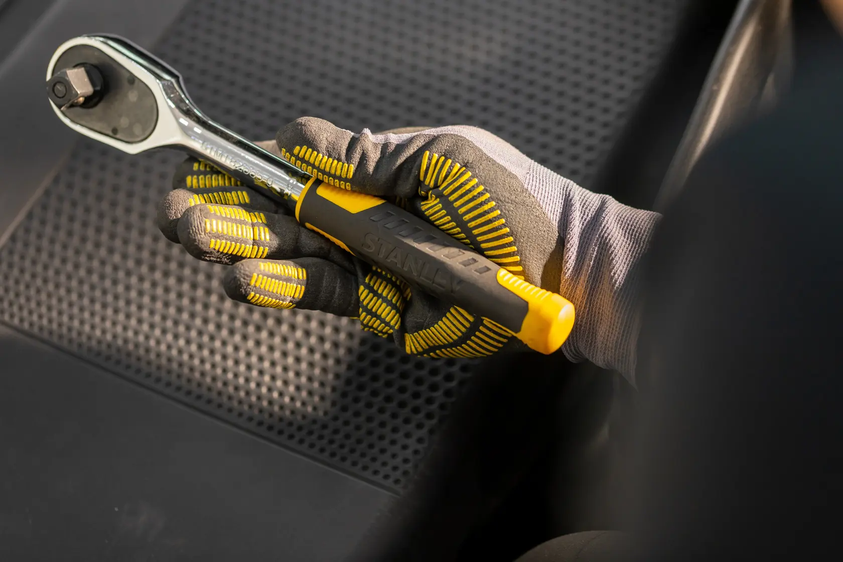 Fotografía de la llave de carraca STANLEY STMT82832-LA con mango amarillo y negro sostenida por una mano enguantada sobre un asiento de automóvil.